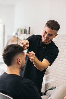 Front view of young barber combing unrecognizable man in barber shop against blurred background - ADSF45716