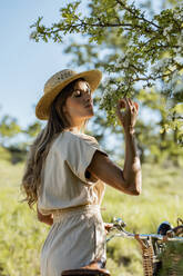 Side view of young gentle female in straw hat and overall standing in countryside with bicycle while smelling blossoming tree during summer weekend - ADSF45729