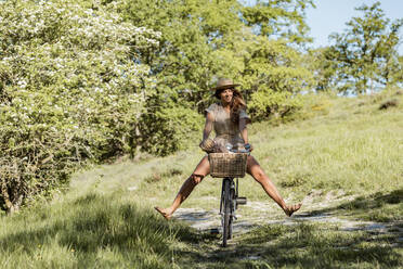 Full body of young positive female in straw hat riding bike with legs up in countryside on sunny summer day having fun time - ADSF45730