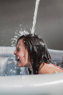 Screaming happy child with wet hair mouth opened and eyes closed sitting in inflatable pool while having fun under falling water from top in sunlight - ADSF45731