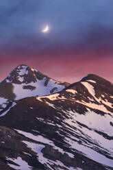 Picturesque view of mountain range with snow against colorful purple sky with glowing curve moon at evening - ADSF45732