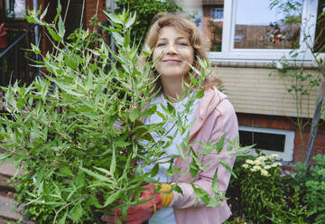 Lächelnde reife Frau mit Salbeipflanze vor einem Haus stehend - IHF01549