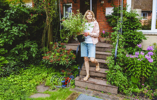 Smiling mature woman moving down on steps with plant - IHF01550