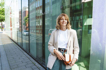 Smiling mature woman standing with sling bag in front of glass wall - IHF01570