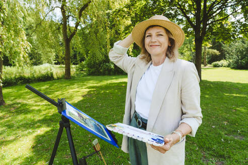 Lächelnde reife Frau mit Hut steht mit Aquarellfarben im Park - IHF01580