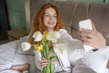 Eine junge Frau fängt einen Moment mit Tulpen in einem Selfie ein, während sie bequem auf ihrem Bett zu Hause sitzt - YBF00005