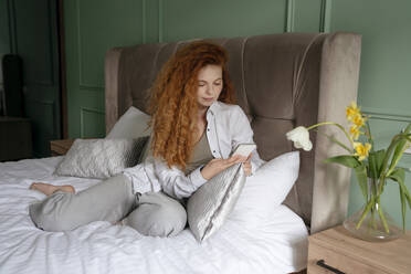 Rothaarige Frau mit Smartphone auf dem Bett sitzend zu Hause - YBF00006