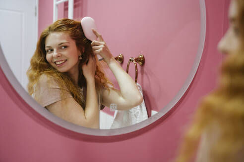 Lächelnde Frau beim Kämmen der Haare im Spiegel zu Hause - YBF00019