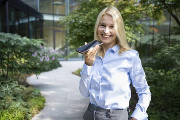 Lächelnde Geschäftsfrau, die eine Sprachnachricht auf einem Mobiltelefon außerhalb des Büros sendet - SVKF01519
