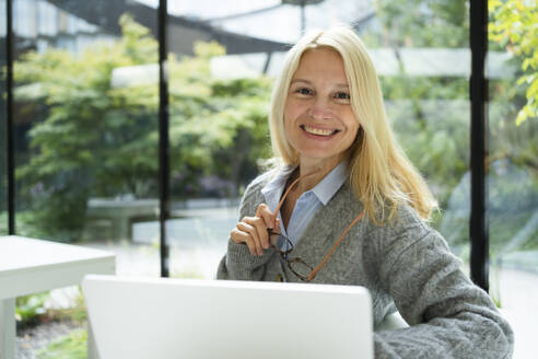Smiling businesswoman holding eyeglasses with laptop at office - SVKF01527