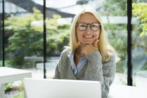 Smiling businesswoman with hand on chin sitting at office - SVKF01528