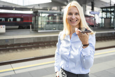 Lächelnde Geschäftsfrau im Gespräch über Lautsprecher am Bahnhof - SVKF01540