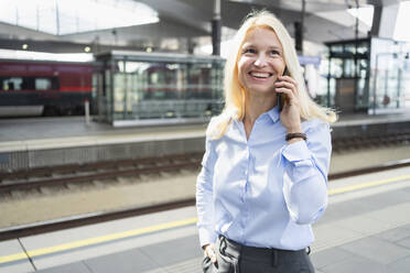 Lächelnde reife Geschäftsfrau, die am Bahnhof mit einem Mobiltelefon spricht - SVKF01541