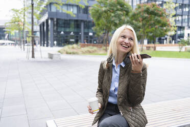 Smiling businesswoman talking on phone and holding disposable cup at office park - SVKF01546