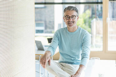 Happy businessman wearing eyeglasses sitting on desk - UUF29577