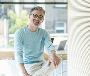 Smiling businessman sitting on desk in office - UUF29578