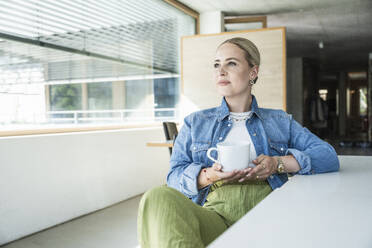 Nachdenkliche junge Geschäftsfrau sitzt mit Kaffeetasse am Schreibtisch - UUF29623