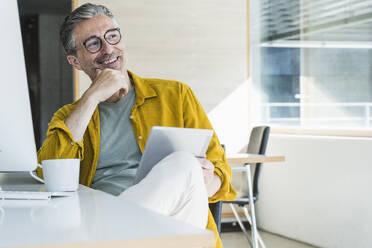 Thoughtful businessman sitting with tablet PC at desk - UUF29632