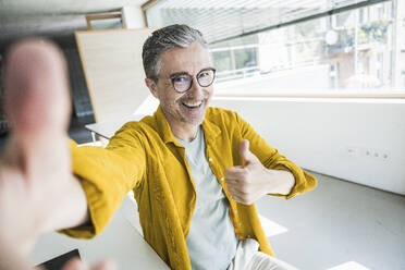 Ein fröhlicher Geschäftsmann zeigt den Daumen hoch und macht ein Selfie in seinem Büro, das Positivität und Erfolg ausstrahlt - UUF29647