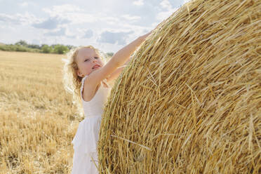 Blondes Mädchen berührt Strohballen auf einem Feld an einem sonnigen Tag - SIF00696