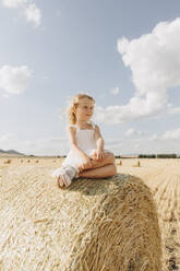 Blondes Mädchen sitzt auf einem Strohballen auf einem Feld an einem sonnigen Tag - SIF00697