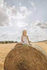 Nachdenkliches blondes Mädchen sitzt auf einem Strohballen auf einem Feld an einem sonnigen Tag - SIF00698