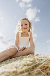 Lächelndes Mädchen sitzt auf einem Strohballen an einem sonnigen Tag - SIF00699
