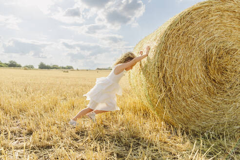 Blond girl pushing bale of straw in stubble field on sunny day - SIF00702