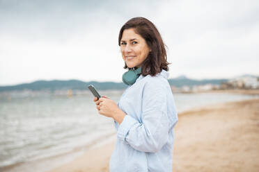 Lächelnde Frau steht mit Smartphone am Strand - JOSEF19987