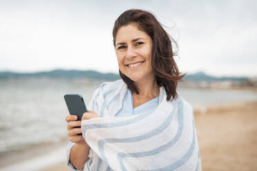 Glückliche Frau mit Smartphone am Strand stehend - JOSEF20010