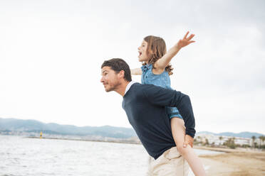 Cheerful girl with father giving piggyback ride at beach - JOSEF20021