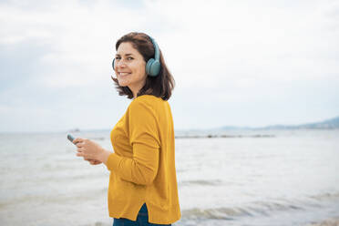 Smiling woman wearing wireless headphones standing with smart phone in front of sea - JOSEF20090