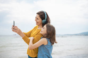 Eine fröhliche Mutter mit kabellosen Kopfhörern macht ein Selfie mit ihrer Tochter mit einem Smartphone - JOSEF20091