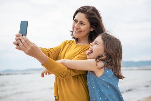 Eine fröhliche Mutter hält einen Moment mit ihrer Tochter am Strand fest und benutzt ihr Smartphone für ein Selfie - JOSEF20092