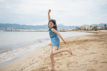 Cheerful girl wearing wireless headphones listening to music and dancing on coastline at beach - JOSEF20096