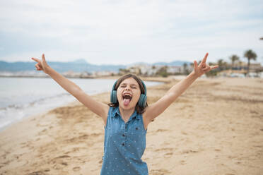 Cheerful girl wearing wireless headphones gesturing horn sign at beach - JOSEF20098