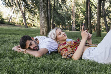 Junge Frau, die ein Smartphone benutzt, mit einem Mann, der sich im Park im Gras entspannt - PBTF00056