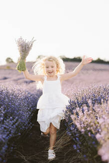 Blondes Mädchen hält einen Strauß Lavendelblüten und läuft in einem Feld - SIF00721