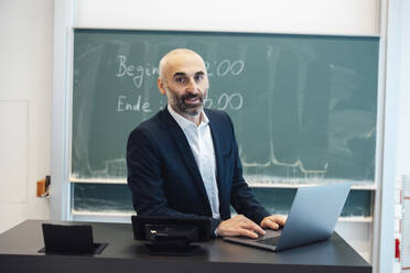 Professor sitting at desk with laptop in lecture hall - JOSEF20167