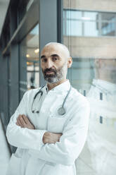 Mature doctor with arms crossed leaning on glass window - JOSEF20175