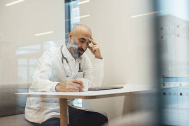 Tired doctor sitting with coffee cup and laptop seen through glass - JOSEF20176