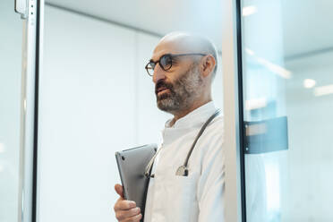 Thoughtful doctor standing with laptop in doorway - JOSEF20207