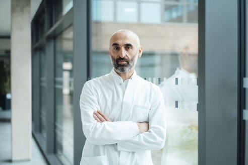 Smiling mature doctor standing with arms crossed by glass - JOSEF20227