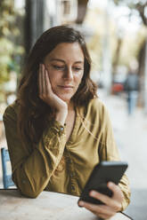 Junge Frau mit Hand am Kinn, die in einem Café ihr Smartphone benutzt - JOSEF20270