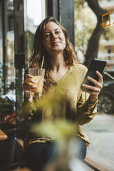 Smiling young woman holding glass of juice and smart phone in cafe - JOSEF20277