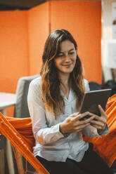 Smiling young businesswoman using tablet PC in hammock in office - JOSEF20309