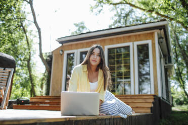 Thoughtful freelancer sitting with laptop in front house - MDOF01418