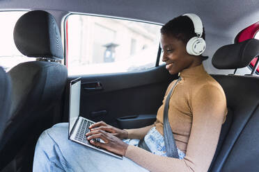 Happy young woman wearing wireless headphones sitting in car - PNAF05765