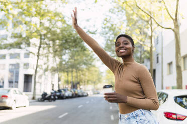 Happy young woman with coffee cup hailing ride on street - PNAF05776