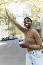 Smiling young woman with coffee cup hailing ride on street - PNAF05777